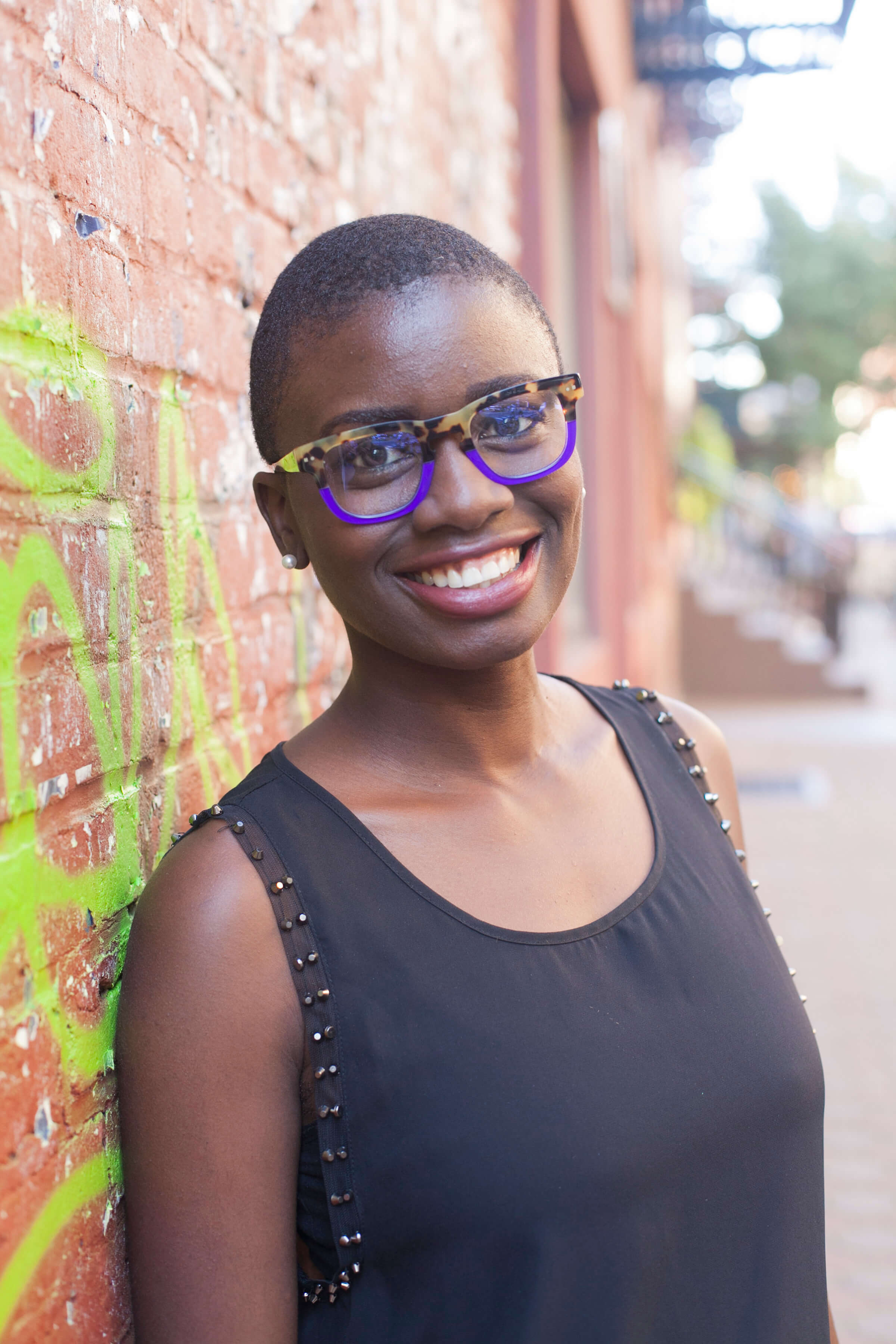 Photo of Sabine Joseph. She is leaning against a brick wall with bright-green graffiti. She is smiling.