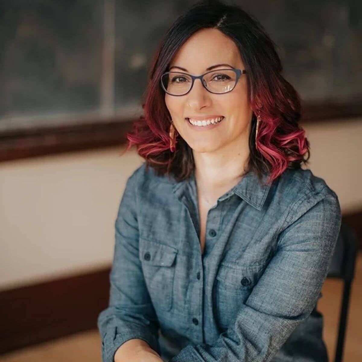 Headshot of artist, illustrator and founder of Polkidots Louma El-Khoury. She turns special outfits into keepsake artworks. Louma has ombre colored shoulder-length hair. It's dark brown on top and dark pink on the bottom. She wears a denim-gray collared button-down and is sitting for this photo. She has brown eyes and wears blue glasses and is smiling. 