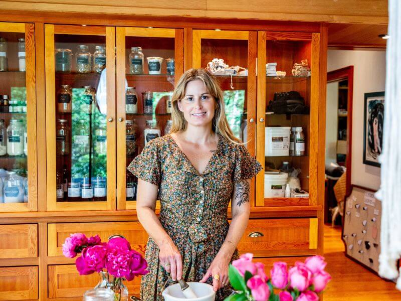 Photo of Meghan MacGregor, owner of Cosmic Skincare. In her interview, Meghan shares how she began selling skincare products online and in boutiques.

In the photo, she is standing in front of apothecary-style cabinets. She is a white woman with long-ish blond hair and is wearing a short sleeve dress with a floral design.