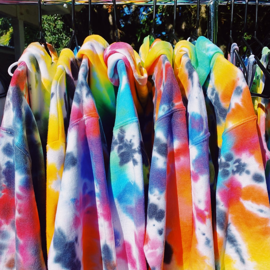 Seven tie-dye sweatshirts hanging on a clothing rack created by Live & Let Dye.