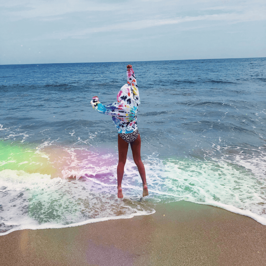 Photo of woman in a bathing suit wearing a tie dye hooded sweatshirt from Let's Get Dye by by Andrea Carneiro. She's jumping at the shore of the ocean.