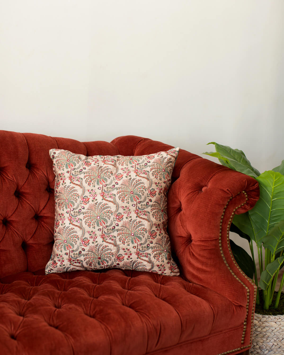 Throw pillow designed by Sushmitha Pidatala of Arjuna Design Studio. The pillow features a green and red floral design and sits on a red tufted sofa. 